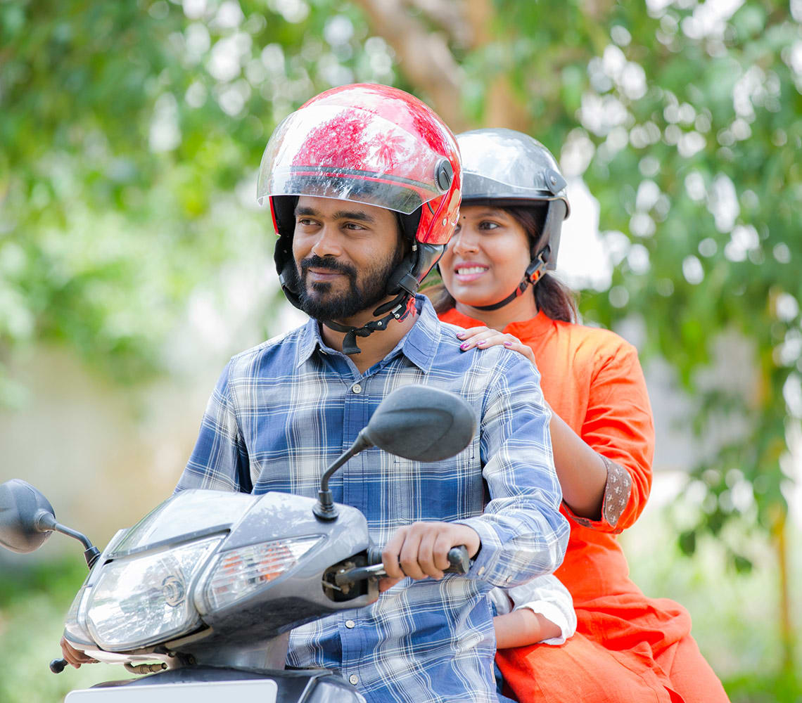 Two people riding a motorbike outdoors, both wearing helmets. The person in front is in a blue plaid shirt and the person in back is in an orange outfit. Trees are visible in the background.